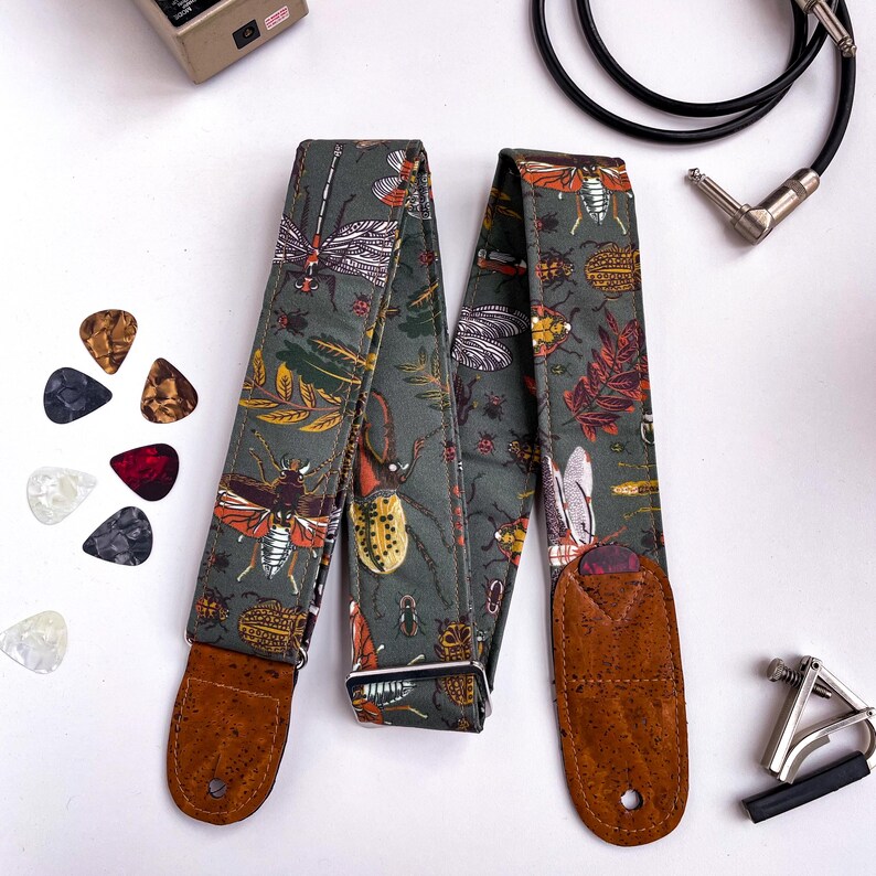 May include: A guitar strap with a dark green background featuring a pattern of insects and leaves in shades of orange, brown, and white. The strap has brown leather ends and a silver metal buckle. Guitar picks and a capo are also visible.