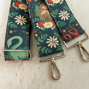 Puede incluir: Tres correas de tela con diferentes patrones florales y un cierre de metal plateado. Las correas son de color azul oscuro con flores blancas y hojas verdes. Una correa tiene una serpiente marina verde, otra tiene un pulpo rojo y la tercera tiene un perezoso marr&oacute;n.