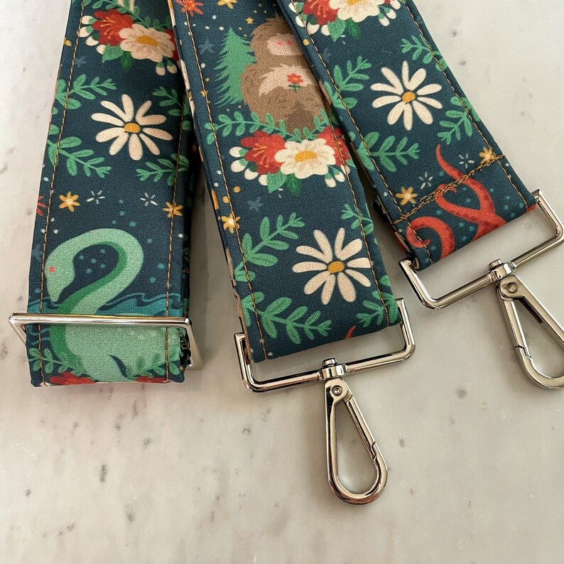 Puede incluir: Tres correas de tela con diferentes patrones florales y un cierre de metal plateado. Las correas son de color azul oscuro con flores blancas y hojas verdes. Una correa tiene una serpiente marina verde, otra tiene un pulpo rojo y la tercera tiene un perezoso marr&oacute;n.