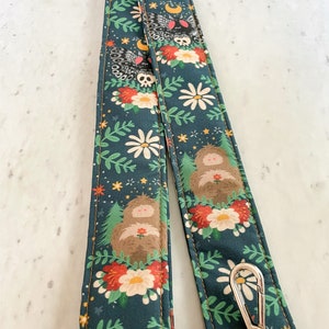 Puede incluir: Una correa de c&aacute;mara verde oscuro con un patr&oacute;n floral que presenta margaritas blancas, flores rojas, hojas verdes y criaturas del bosque marrones. La correa tiene un cierre de metal plateado.