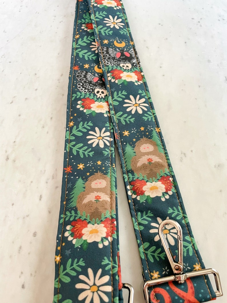 Puede incluir: Una correa de c&aacute;mara verde oscuro con un patr&oacute;n floral que presenta margaritas blancas, flores rojas, hojas verdes y criaturas del bosque marrones. La correa tiene un cierre de metal plateado.