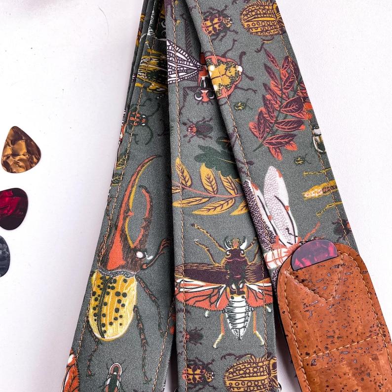 May include: A guitar strap with a gray background and a repeating pattern of colorful insects and leaves. The strap has a brown cork end and is paired with several guitar picks.