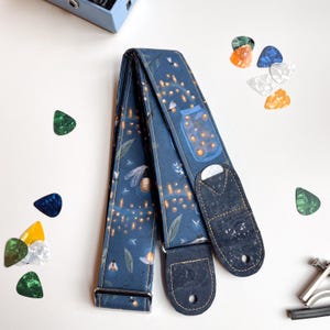 May include: A blue guitar strap with a firefly and jar design. The strap has cork ends and a silver buckle.