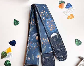 Puede incluir: Una correa de guitarra azul con un diseño de luciérnagas y tarros. La correa tiene extremos de corcho y una hebilla plateada.