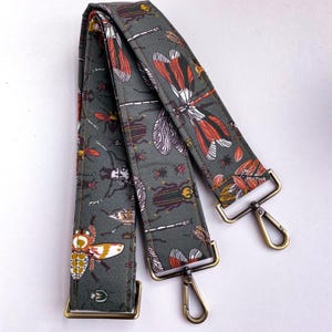 May include: A dark green patterned bag strap featuring various insects, including dragonflies and beetles, in shades of orange, white, and brown. The strap has gold-colored metal clasps and rectangular loops for attachment.