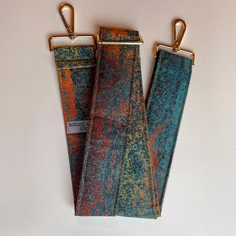 May include: A long, adjustable, multi-colored fabric strap with gold hardware. The strap has a blue, orange, and yellow abstract pattern. The strap is labeled "Coco Straps".