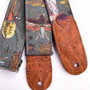 May include: A guitar strap with a gray background and a pattern of various insects in orange, yellow, and white. The strap has brown cork ends and a guitar pick holder.