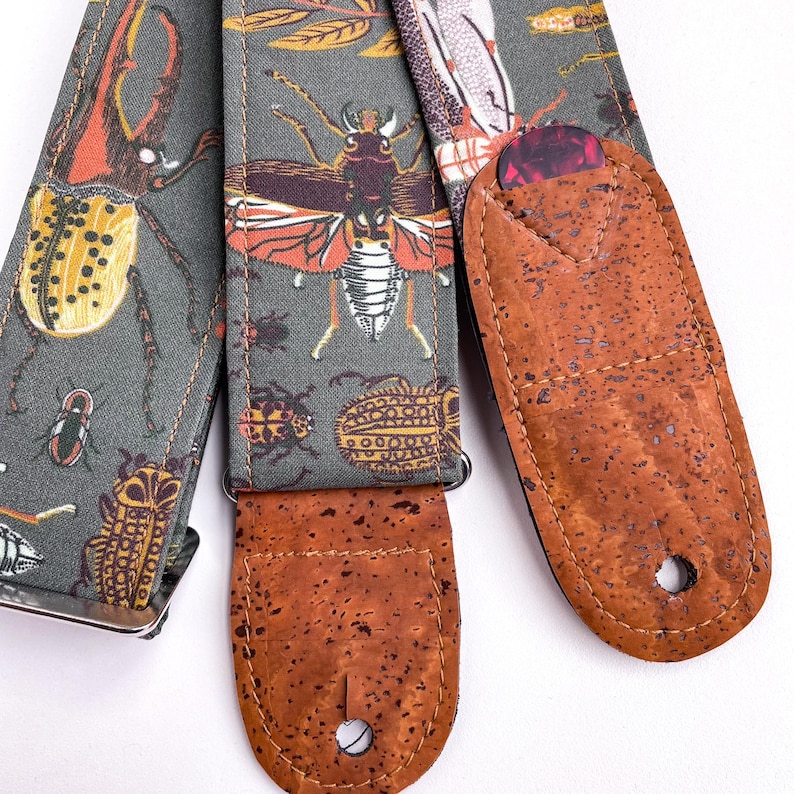 May include: A guitar strap with a gray background and a pattern of various insects in orange, yellow, and white. The strap has brown cork ends and a guitar pick holder.