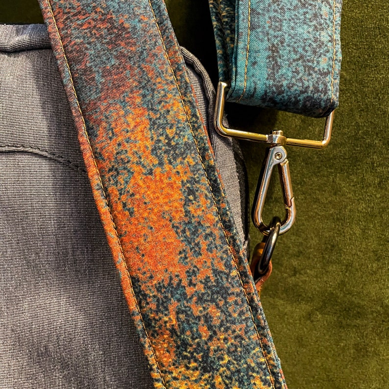 May include: A close-up of a brown and blue patterned fabric strap with a gold metal clasp. The strap is attached to a gray fabric bag.