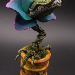 Può includere: Una scultura stravagante di una pianta carnivora con una grande bocca dentata e vibranti petali blu e viola. La pianta è in un vaso giallo e arancione con un simbolo di radiazione.