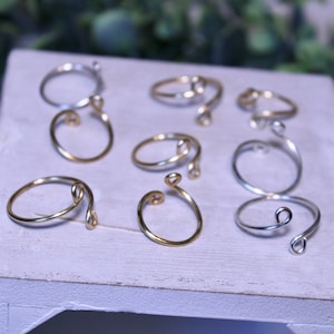 May include: A collection of eight silver and gold wire rings with a unique twist design. The rings are arranged on a white wooden surface.
