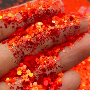 May include: Close-up of bright orange glitter with a holographic finish. The glitter is scattered on a hand, showing its texture and sparkle.