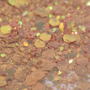 May include: Close-up of a pile of iridescent glitter in shades of peach, gold, and pale green. The glitter particles are hexagonal and of varying sizes, creating a textured, sparkling effect. The overall aesthetic is soft and luminous.