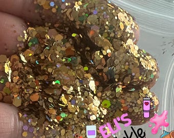 May include: A close-up of a large amount of gold glitter with iridescent flecks. The glitter is in a clear plastic bag and is being held by a hand. The text "ELI'S Twinkle DUST" is visible in the image.