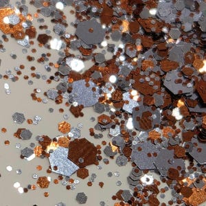 May include: Close-up of a scattering of metallic glitter in various sizes and shapes. The glitter is primarily silver and copper-colored, with a few specks of white. The glitter is scattered on a light beige surface.