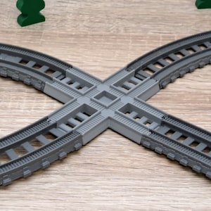 May include: A gray plastic toy train track set with four curved pieces and a cross-shaped connector. The track is designed to connect to other pieces of track.