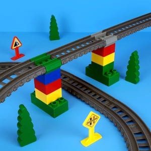 May include: A toy train track set featuring a bridge supported by colourful blocks. The set includes grey tracks, green trees, and yellow road signs against a blue backdrop. The bridge is supported by red, yellow, green, and blue blocks.