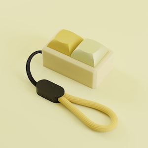 May include: A two-tone yellow and light green keyboard keycap set with a black wrist strap. The keycaps are square and sit in a rectangular base. The wrist strap is yellow and black.
