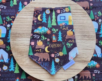 May include: A brown bandana with a camping theme. The bandana features a repeating pattern of trees, campfires, tents, and a camper van. The text "Explore" and "Camp" are also included in the pattern.