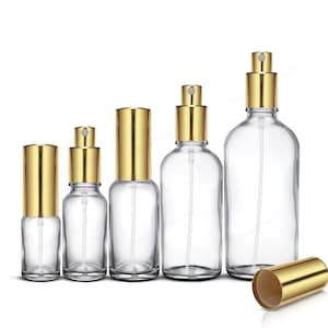 May include: A selection of clear glass spray bottles with gold-coloured metal tops and spray mechanisms. The bottles are of varying sizes, with one gold cap lying on its side. The bottles are designed for dispensing liquids.