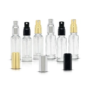 May include: Six clear glass bottles with gold, silver and black spray tops. The bottles are empty and are arranged in a row. There are three additional spray tops in the foreground, one gold, one silver and one black.