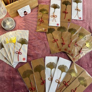 May include: A collection of ginkgo leaf bookmarks. Each bookmark features a pressed ginkgo leaf with a red or gold-colored string bow. The bookmarks are packaged on white and brown cardstock.