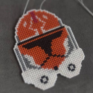 May include: Cross-stitch of a Star Wars clone trooper helmet in orange, black and white.
