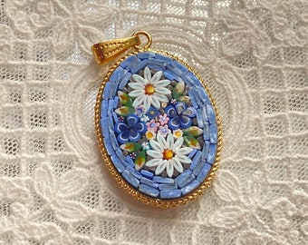 Puede incluir: Un colgante ovalado de oro con un diseño de mosaico azul y blanco con un patrón floral. El colgante tiene una anilla de oro.