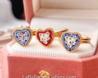 May include: Three gold rings with heart-shaped mosaic designs in a pink jewelry box. Each ring features a different color scheme: blue with pink flowers, red with white flowers, and blue with white flowers. The website address LaBallerinaGallery.com is visible.