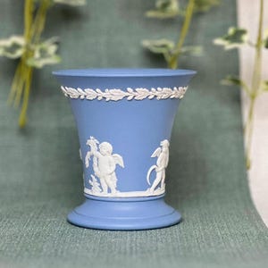 May include: A light blue ceramic vase with a white floral design and cherub figures. The vase has a flared top and a round base.