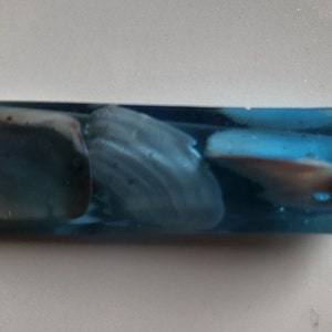 May include: A rectangular pendant with a light blue resin base, encasing three seashells. The shells are white and tan, and the resin has a slight shimmer. A silver-colored metal loop is attached for hanging.