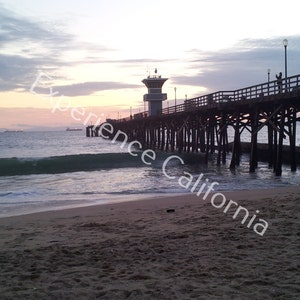 May include: A coastal scene featuring a wooden pier stretching out into the sea at twilight. The sky displays hues of blue and pink, with figures on the pier. The beach and waves are in the foreground.