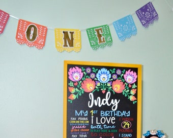 May include: A chalkboard sign with a colorful floral border and the text "Indy My 1st Birthday I Love bath time I weigh 24 pounds to play hide & seek to color I stand 29 tall I have 8 teeth fav foods corn on the cob blue berries juice grilled cheese boiled eggs fav toys piano fluffy toys kitchen toys nicknames monkey monster Indy Snow sunflower give high fives I can clap wave bye". A small doll wearing a pink dress is standing next to the sign.