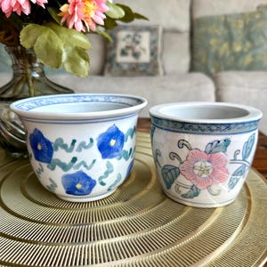 May include: Two small, white ceramic pots with blue and pink floral designs. One pot features blue morning glories, while the other has pink flowers and green leaves. Both have blue rims and sit on a gold-coloured, circular tray.