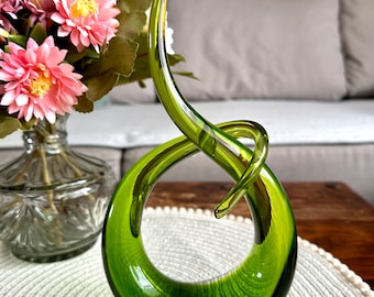May include: A vibrant green glass sculpture with a looped design. The art piece has a smooth, glossy finish and sits on a white placemat. A vase of pink and yellow flowers is in the background.