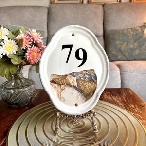 May include: An oval white ceramic house number plaque featuring the black number 79 and a detailed illustration of a bird feeding chicks in a nest. The plaque has a distressed edge and two mounting holes, displayed on a decorative metal stand.
