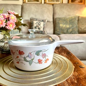 Op de afbeelding: Een witte keramische steelpan met een lange steel en een doorzichtig glazen deksel. De buitenkant van de pan is voorzien van een decoratief bloemmotief met roze, oranje en groene pronkerwtbloemen en ranken rondom de zijkant van de pan.