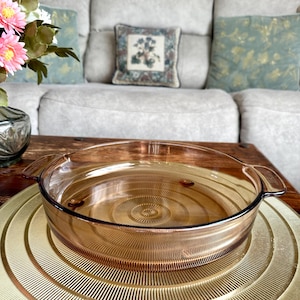 Könnte beinhalten: Eine transparente, bernsteinfarbene Glasbackform mit Griffen, die auf einem goldfarbenen, kreisförmigen Serviertablett steht. Das Tablett hat ein konzentrisches, geriffeltes Design. Der Hintergrund umfasst ein graues Sofa mit dekorativen Kissen und einen Holztisch.