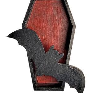 May include: A black wooden bat silhouette emerging from a red wooden coffin. The coffin is framed in black wood.