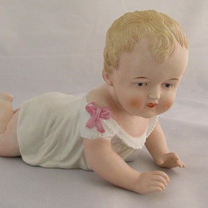 May include: A porcelain baby doll figurine in a white dress with a pink bow. The doll is lying on its stomach with its arms outstretched and its head turned to the side.