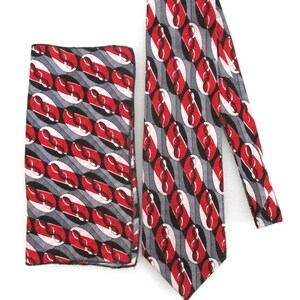May include: A matching tie and pocket square set. The tie and pocket square feature a repeating pattern of red, white, and black oval shapes on a gray background. The tie is displayed next to the folded pocket square.