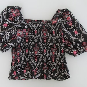 May include: A black blouse featuring a square neckline, short puff sleeves, and a smocked bodice. The fabric is decorated with a repeating white and pink floral damask pattern, and the garment is finished with a ruffled hem at the waistline.