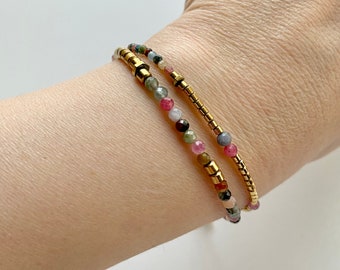 May include: Two bracelets with gold beads and colorful gemstone beads. The bracelets are stacked on a person's wrist.
