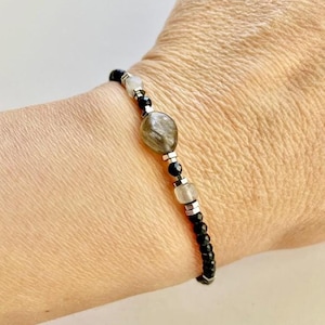 May include: A beaded bracelet featuring a central, oval-shaped, gray-green stone. The bracelet includes black and clear beads, with silver-colored metal accents. The bracelet is worn on a wrist.