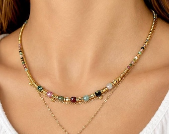 Könnte beinhalten: Eine geschichtete Goldhalskette mit zwei verschiedenen Ketten. Die obere Kette ist mit kleinen, mehrfarbigen facettierten Edelsteinperlen verziert, die mit goldenen zylindrischen Abstandshaltern durchsetzt sind. Die untere Kette ist ein einfaches Glied.