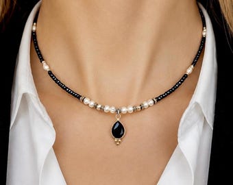 Puede incluir: Un collar con un colgante central de piedra negra en forma de lágrima, suspendido de una hilera de pequeñas cuentas negras intercaladas con perlas blancas y espaciadores metálicos, creando un contraste equilibrado entre tonos oscuros y claros.
