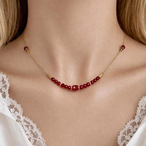 Op de afbeelding: Een delicate gouden ketting met kleine, ronde, dieprode kralen afgewisseld met gouden accenten. De ketting heeft een centrale rechthoekige rode kraal. De ketting wordt gedragen.