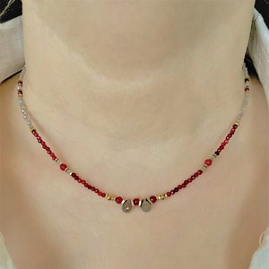 May include: A delicate necklace featuring red and grey beads with two silver teardrop pendants.
