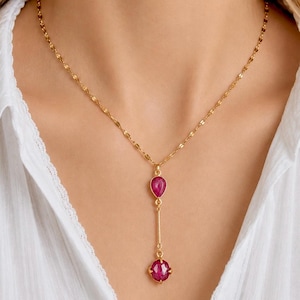 Può includere: Una collana color oro con un pendente con due pietre preziose rosa. La gemma superiore è a forma di goccia, e quella inferiore è rotonda con un'incastonatura decorativa. La collana è indossata su una camicia bianca.