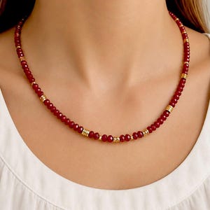 Op de afbeelding: Een ketting met kleine, gefacetteerde, donkerrode kralen gerangschikt in een doorlopende streng. Het ontwerp wordt op regelmatige afstanden onderbroken door setjes van drie dunne, gepolijste goudkleurige metalen ringetjes en enkele goudkleurige tussenkralen.